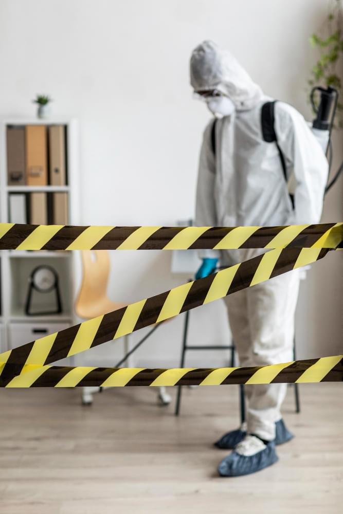 Pracownik w kombinezonie ochronnym przeprowadza dezynfekcję pomieszczenia, za taśmą ostrzegawczą.