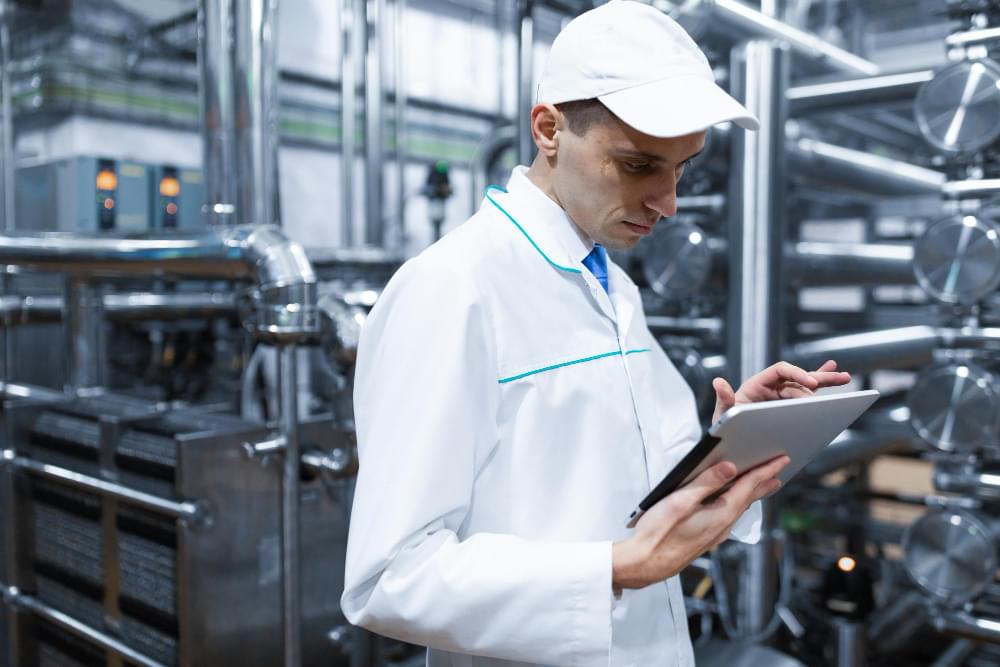 Audytor w białym fartuchu ochronnym i czapce z daszkiem wprowadza dane do tabletu na hali produkcyjnej pełnej stalowych instalacji, nadzorując rygorystyczne procedury systemu HACCP.